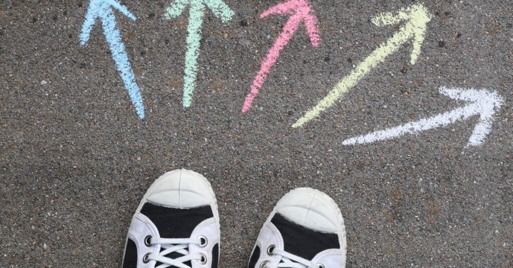 Black sneakers on pavement with colorful chalk arrows pointing in different directions.