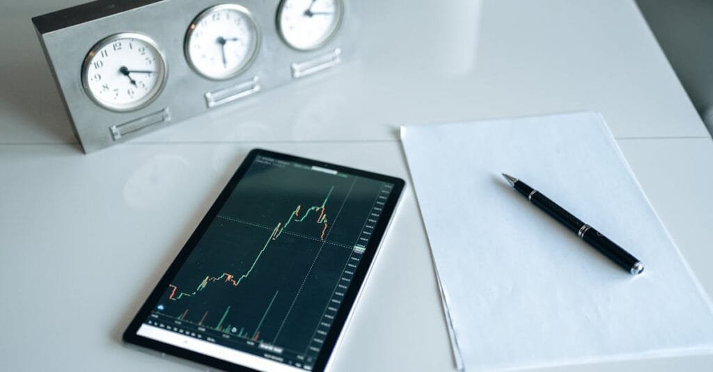 Tablet displaying stock chart on a minimal white workspace with clock and pen.