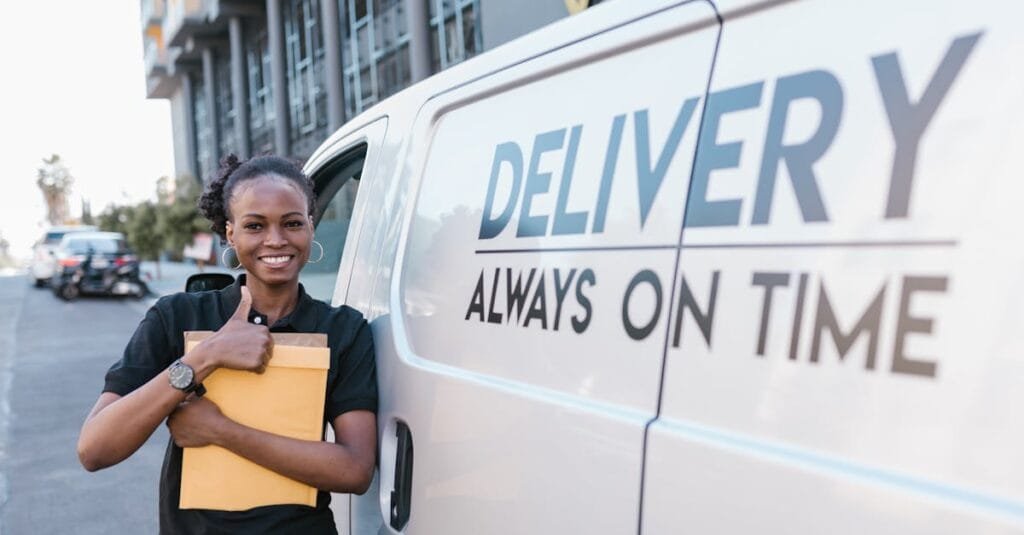 A cheerful delivery woman standing by a van with a parcel, ready for delivery.