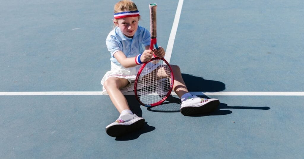 A young girl having fun sitting on a tennis court with a racket.