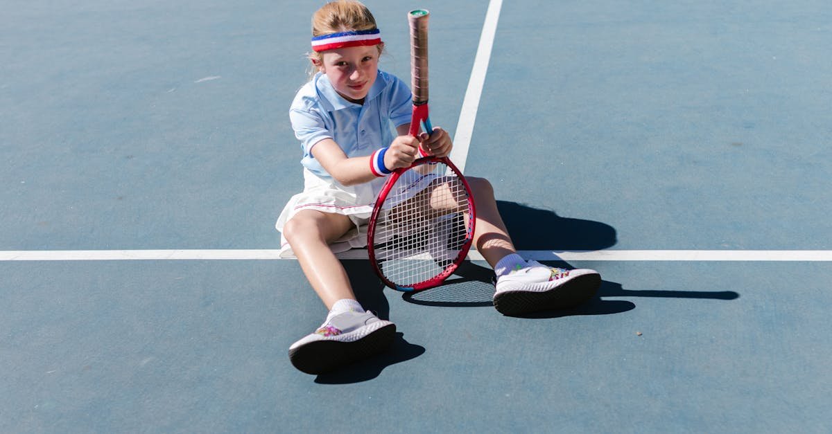 A young girl having fun sitting on a tennis court with a racket.