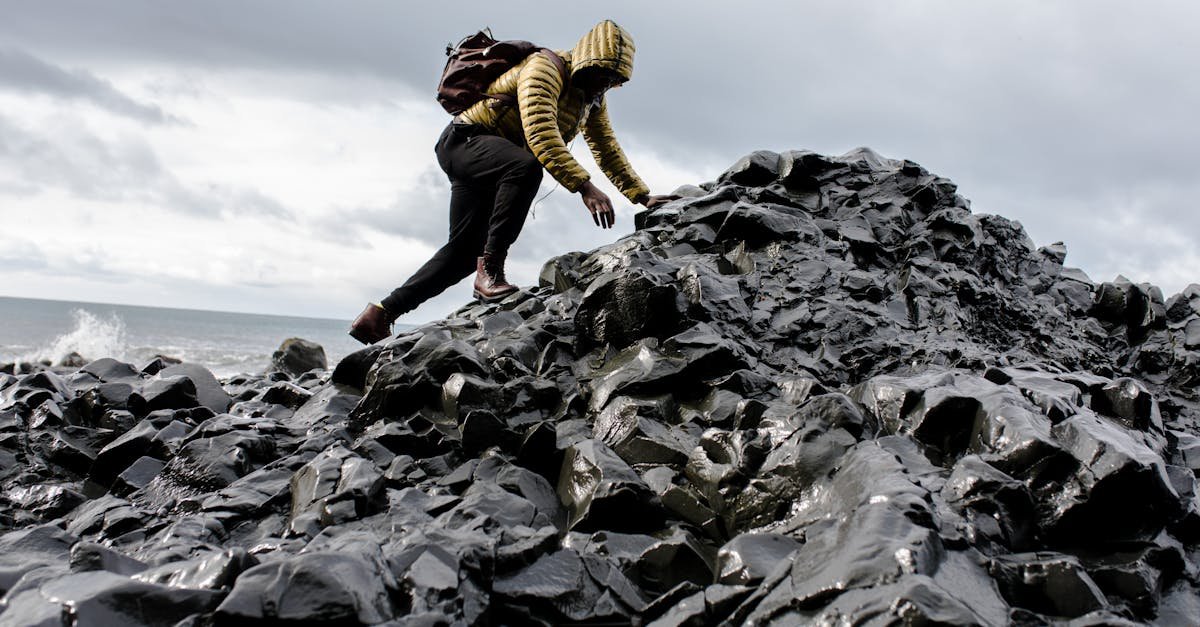 Person in yellow jacket climbs black rocky terrain by the ocean under cloudy skies.