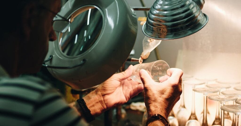 An artisan carefully shaping a glass piece under magnifying lamp indoors.