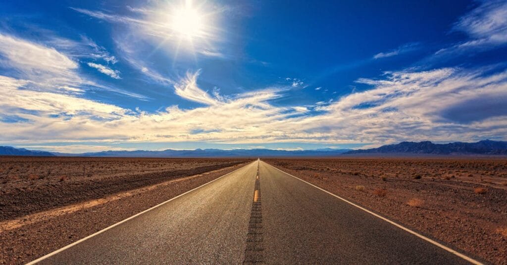 A long, straight road stretches across a dry desert landscape under a bright blue sky and shining sun.