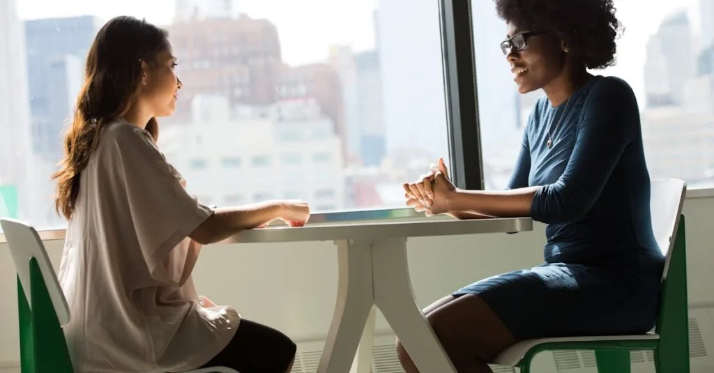 Two women sitting and talking at a table with a city view from the window.