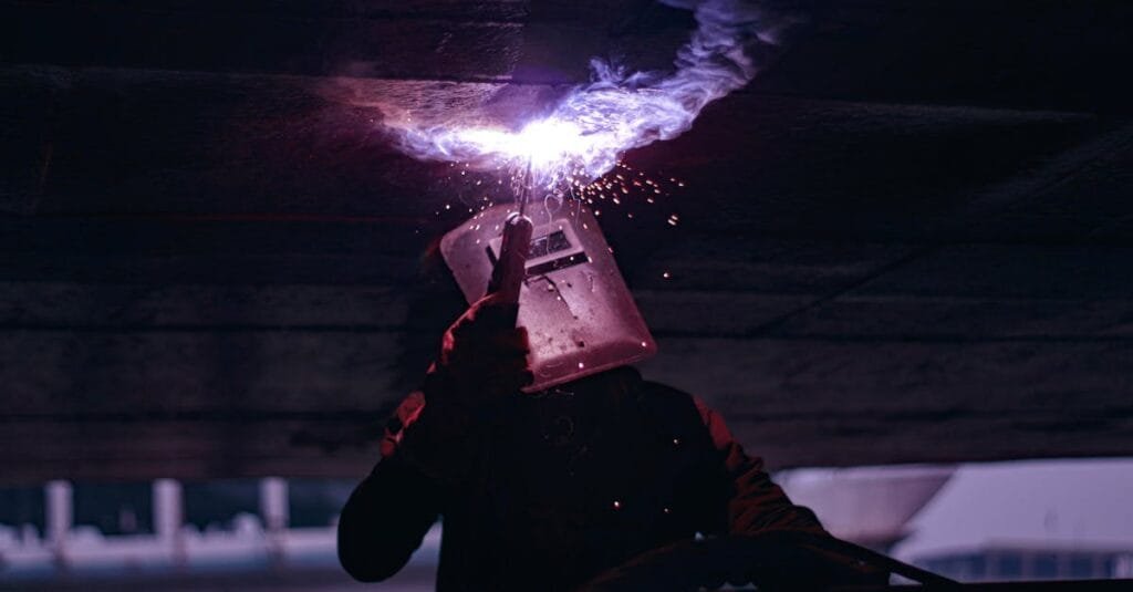 Silhouetted welder working under large metal structure, sparks flying, industrial setting.