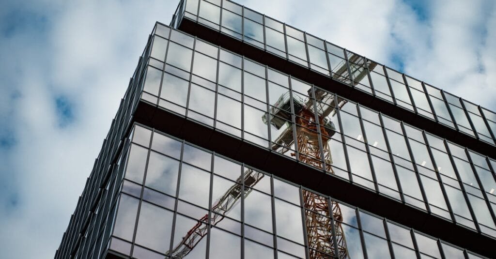 A low angle view of a modern glass building reflecting a construction crane against a cloudy sky.