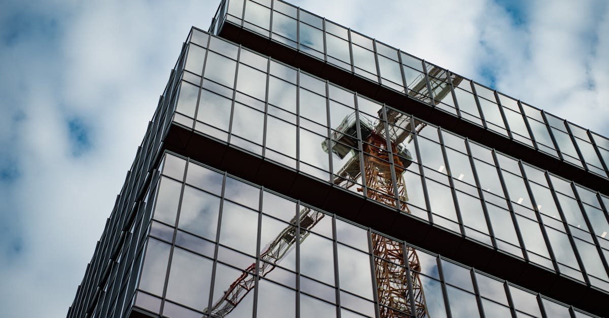 A low angle view of a modern glass building reflecting a construction crane against a cloudy sky.