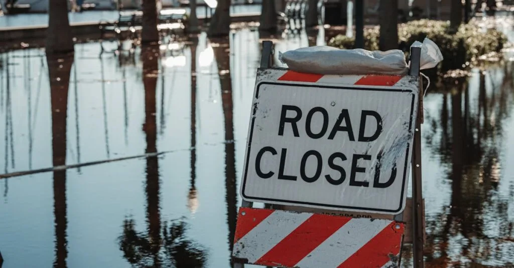 Road closed sign amidst flooded street, reflecting calm water and trees.