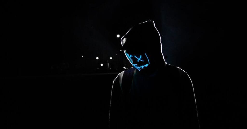 Silhouette of a person in a hoodie wearing a glowing LED mask against a dark background.