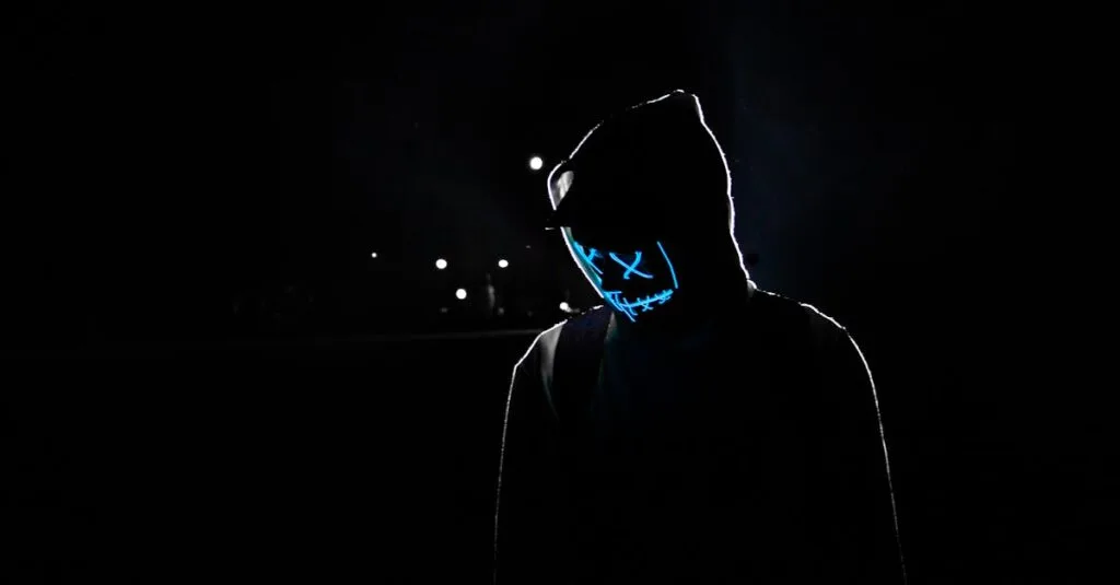 Silhouette of a person in a hoodie wearing a glowing LED mask against a dark background.