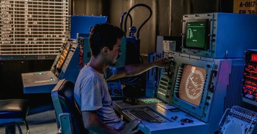 A man working at control panels in a dimly lit industrial room with various monitors.
