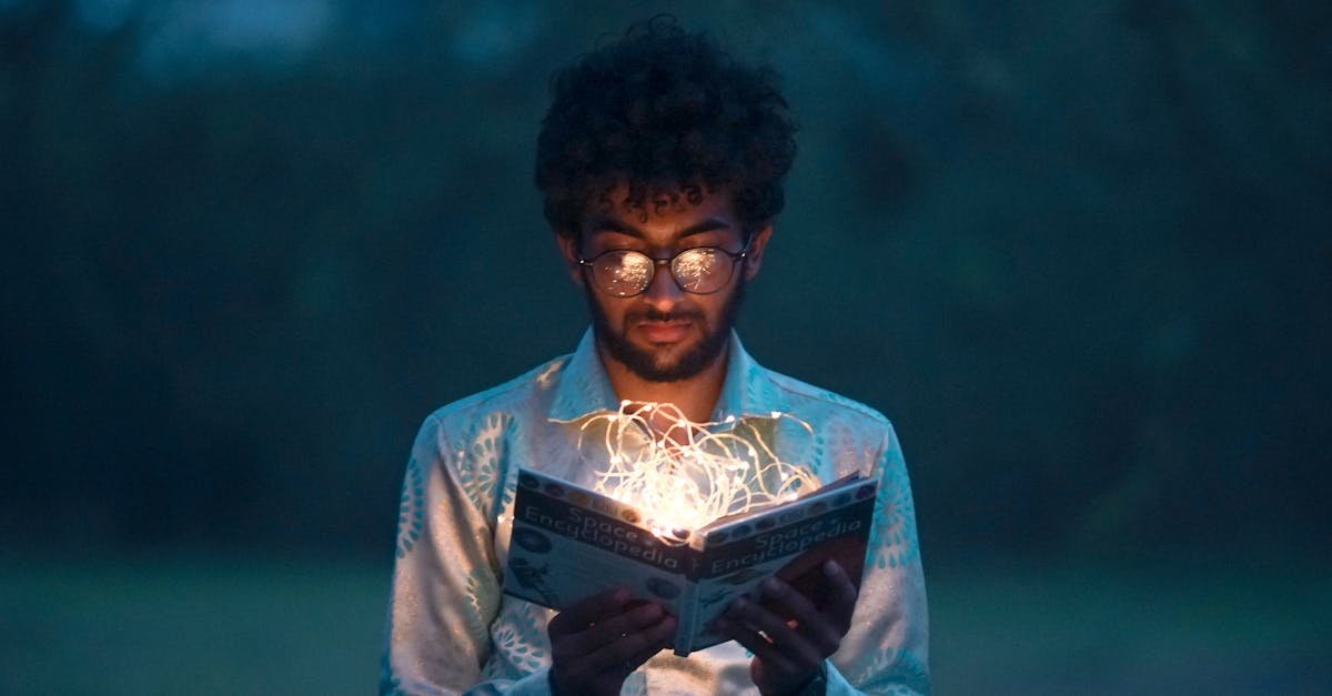A man with glasses reads an illuminated book outdoors during twilight.