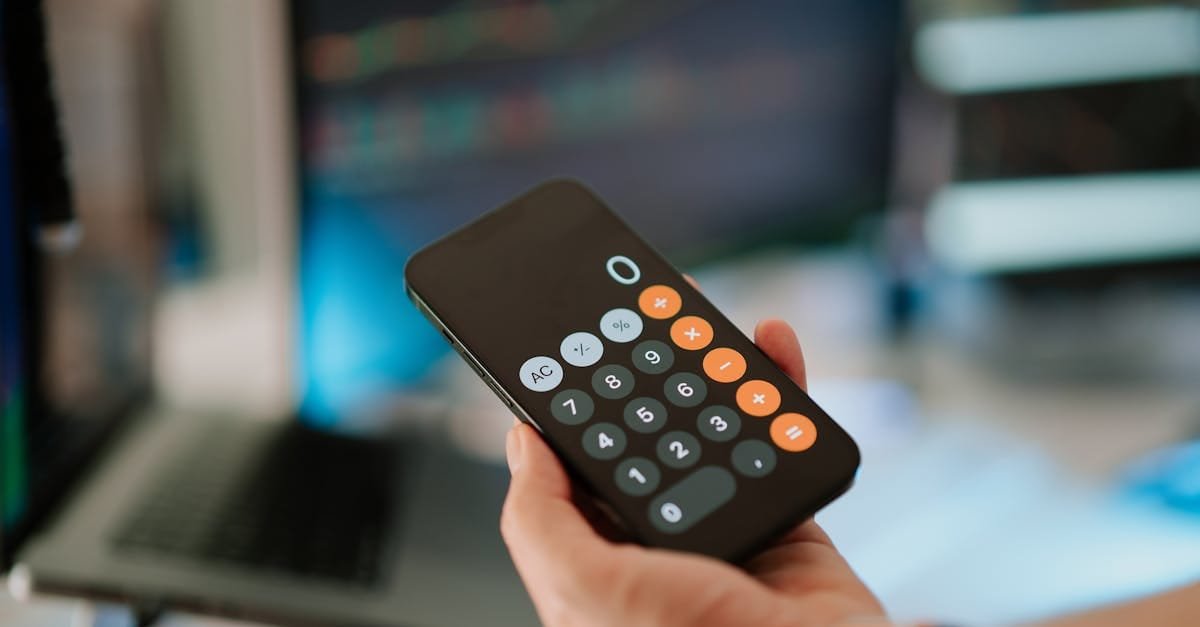 Close-up of hand holding smartphone calculator in office setting with financial data on screen.