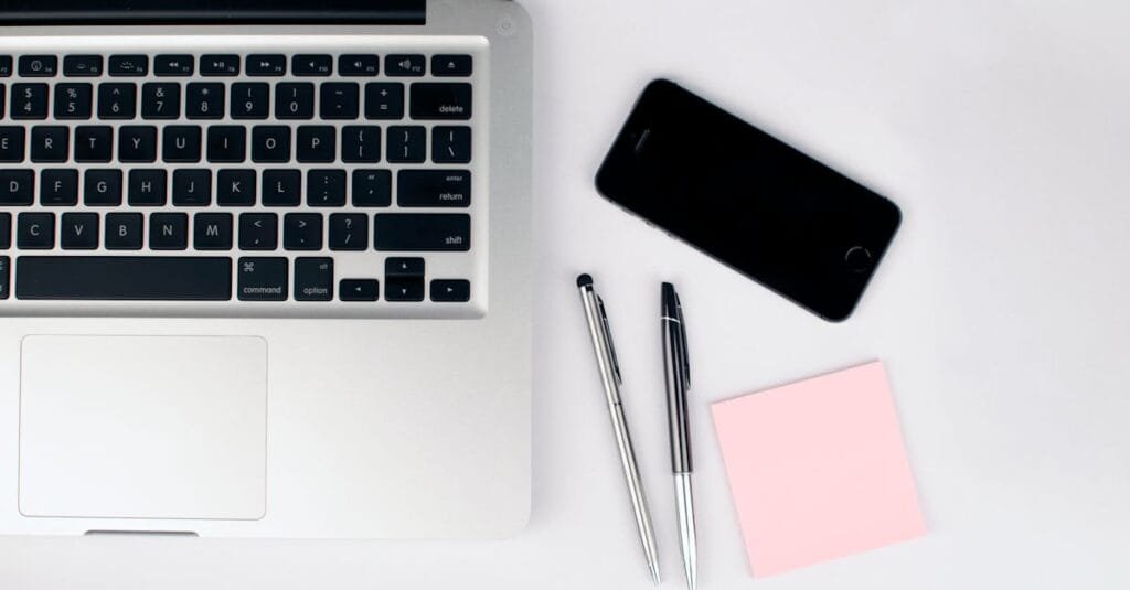A top view of a modern workspace featuring a laptop, smartphone, pens, and a pink sticky note on a white desk.