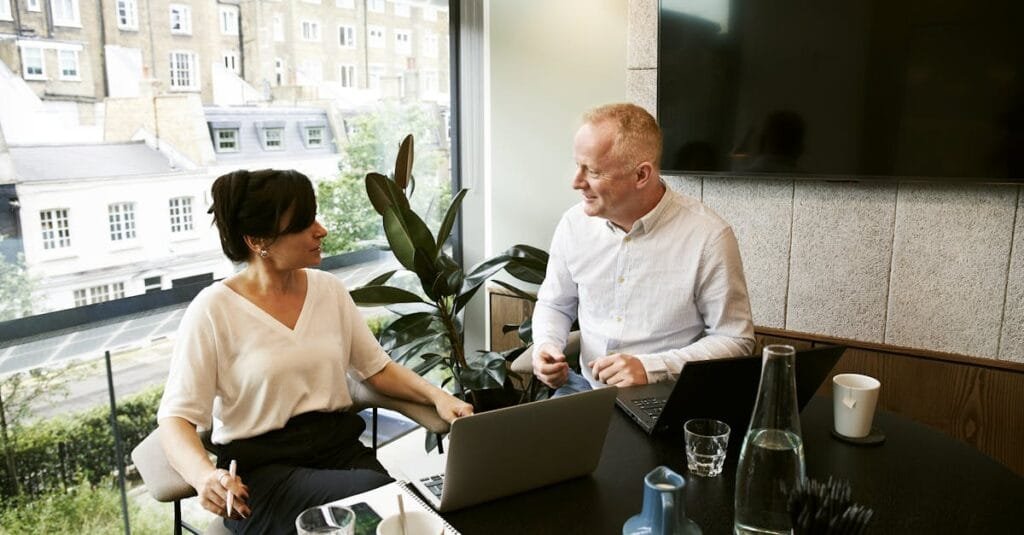 Two business professionals engaging in a team meeting in a modern London office with a large window view.