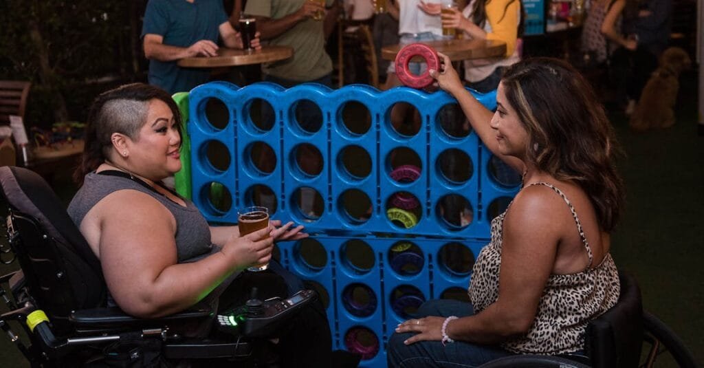 Two women in wheelchairs playing connect four at an indoor bar setting, enjoying a lively social atmosphere.