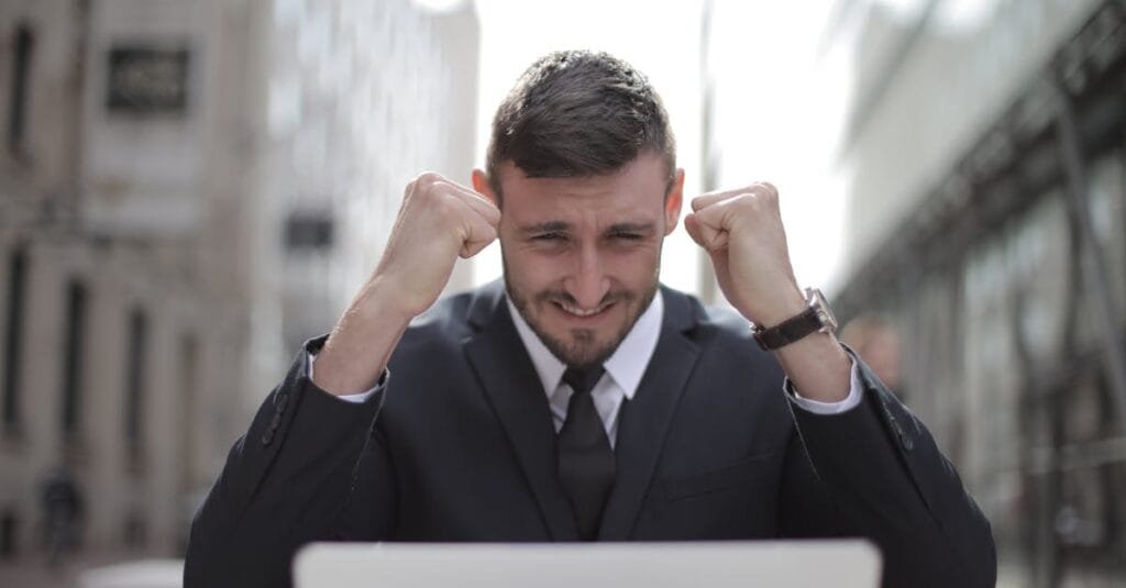 A joyful businessman in a suit triumphantly celebrates success on his laptop outdoors.