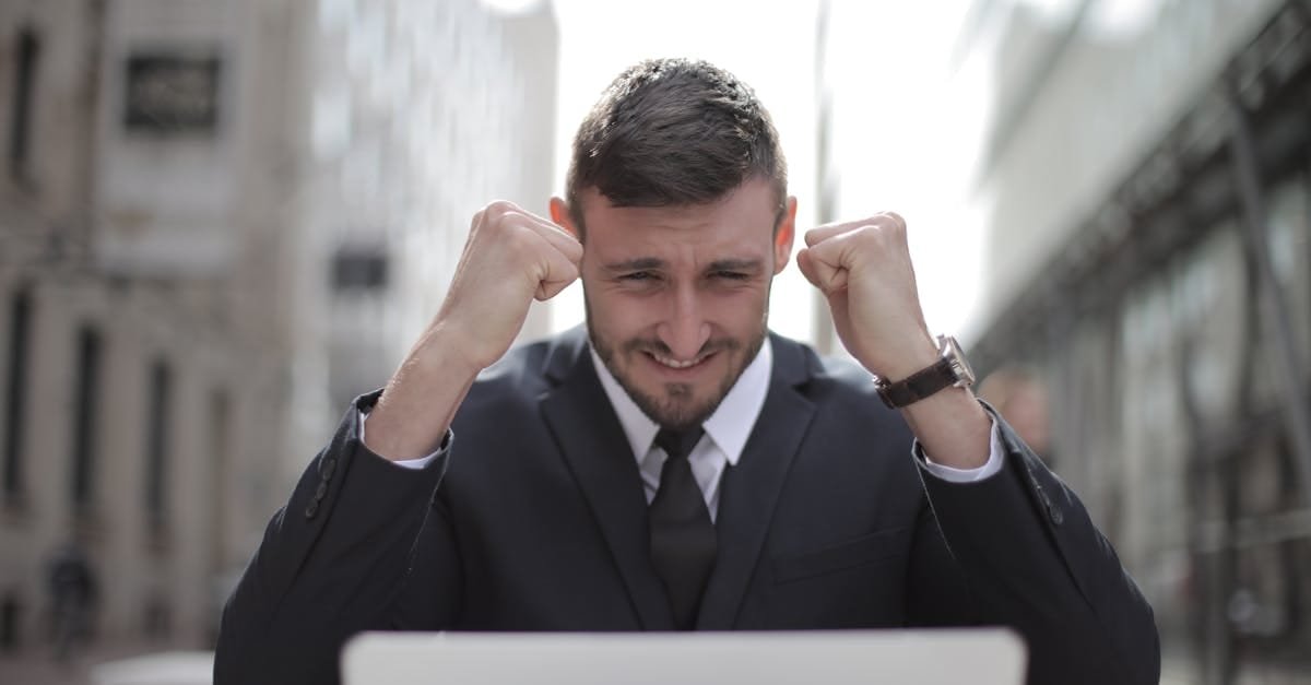 A joyful businessman in a suit triumphantly celebrates success on his laptop outdoors.