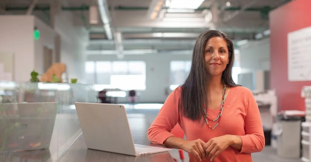 Smiling woman in a bright office, standing by her laptop, exuding confidence.