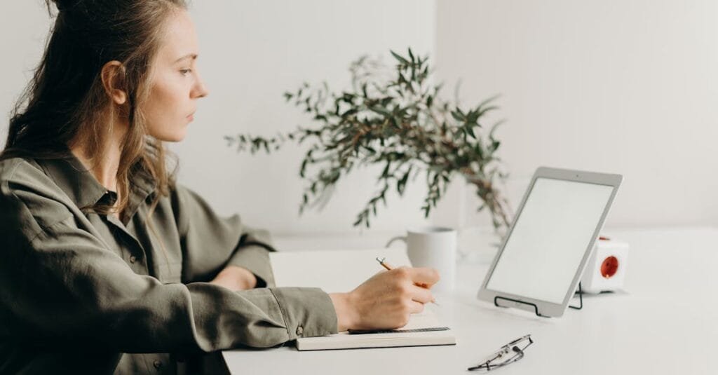 Freelancer writing notes in a minimalist home office with a white tablet, plant, and reading glasses.