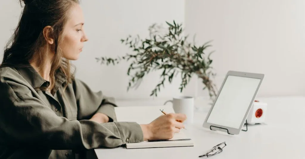 Freelancer writing notes in a minimalist home office with a white tablet, plant, and reading glasses.