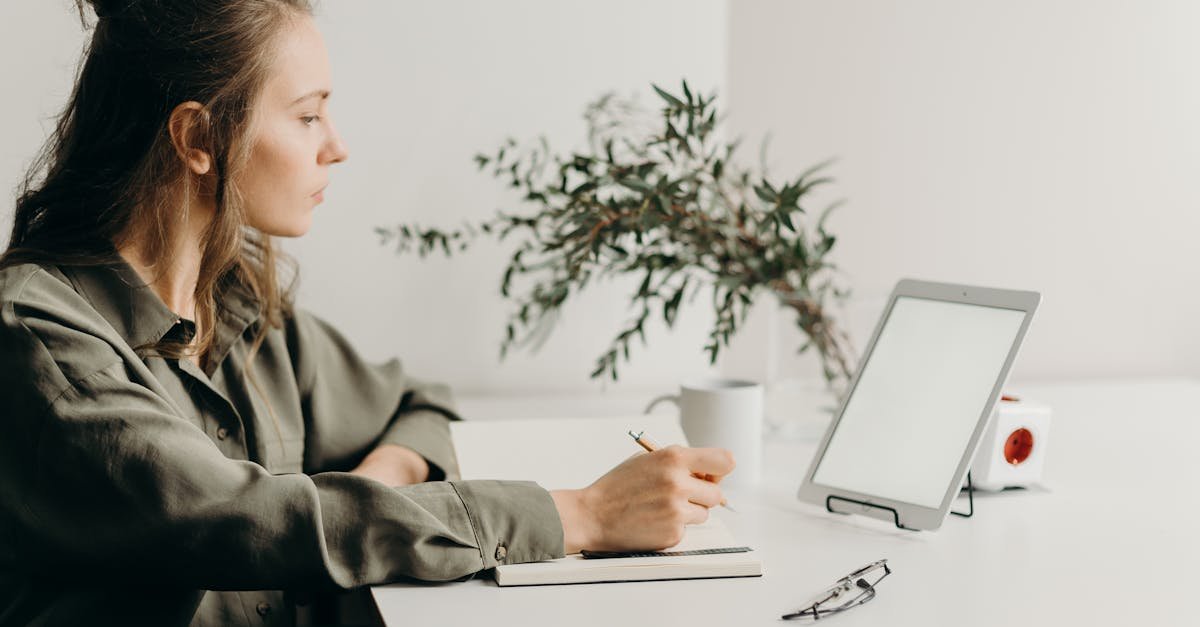 Freelancer writing notes in a minimalist home office with a white tablet, plant, and reading glasses.