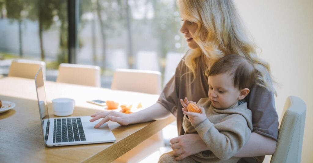 A mother works on a laptop at home while holding her baby, showcasing remote working and parenting.