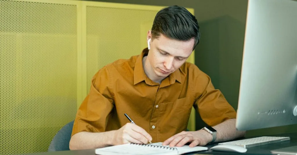 A man concentrating on his studies, taking notes at a desk with a computer.