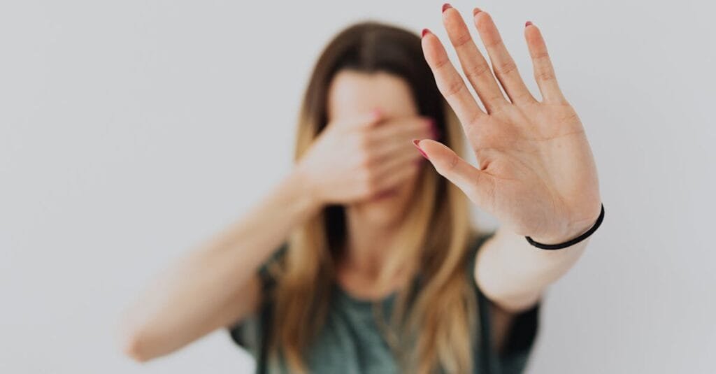 A woman in a gray shirt covers her face with her hand in a stop gesture, evoking a sense of fear and protection.