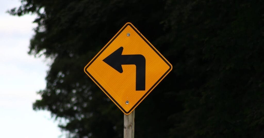 Close-up of a yellow left turn warning sign on a rural road.