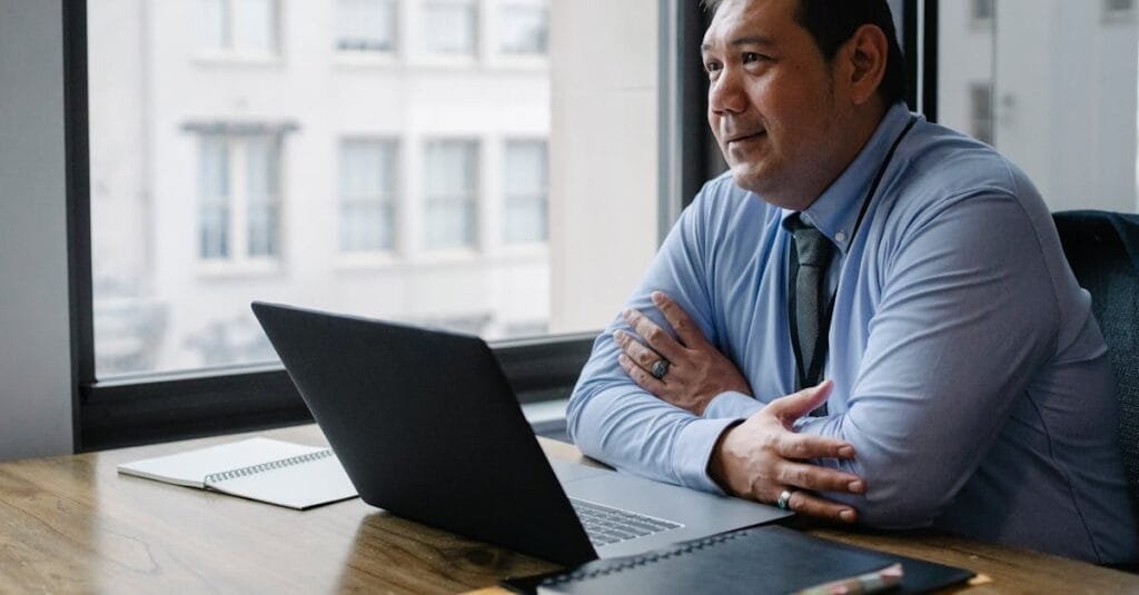 Adult successful ethnic male boss wearing shirt and tie sitting with hands crossed at workplace with documents and netbook
