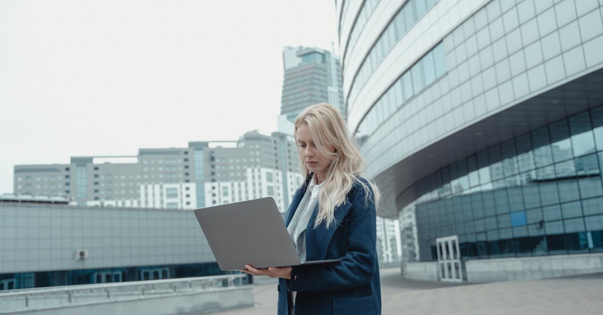Blonde woman in a blue coat using a laptop outside a modern urban building.