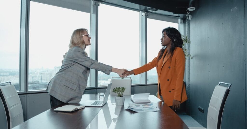 Two businesswomen shaking hands in a modern office setting, symbolizing partnership and agreement.
