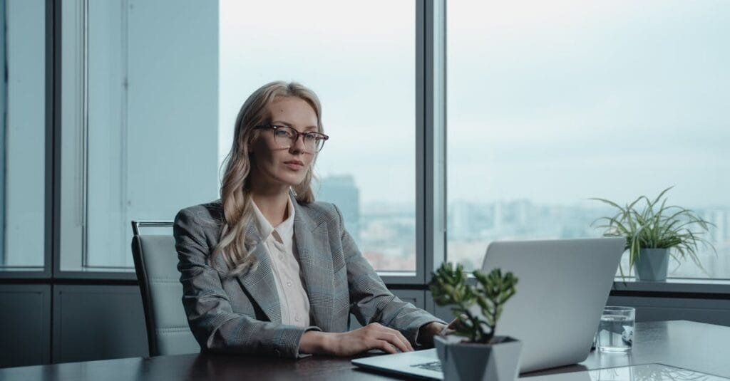 A confident businesswoman in a grey suit working on a laptop in a modern office setting.
