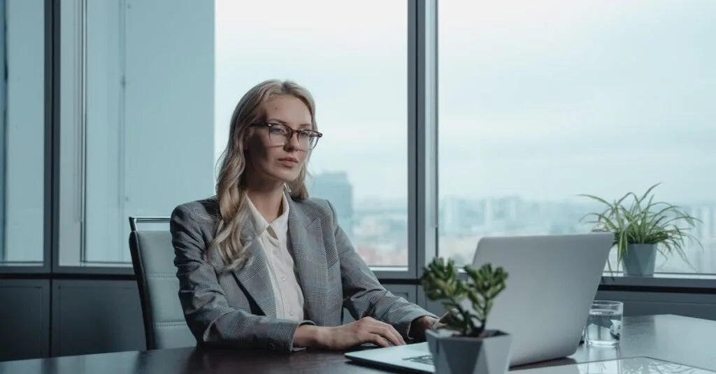 A confident businesswoman in a grey suit working on a laptop in a modern office setting.