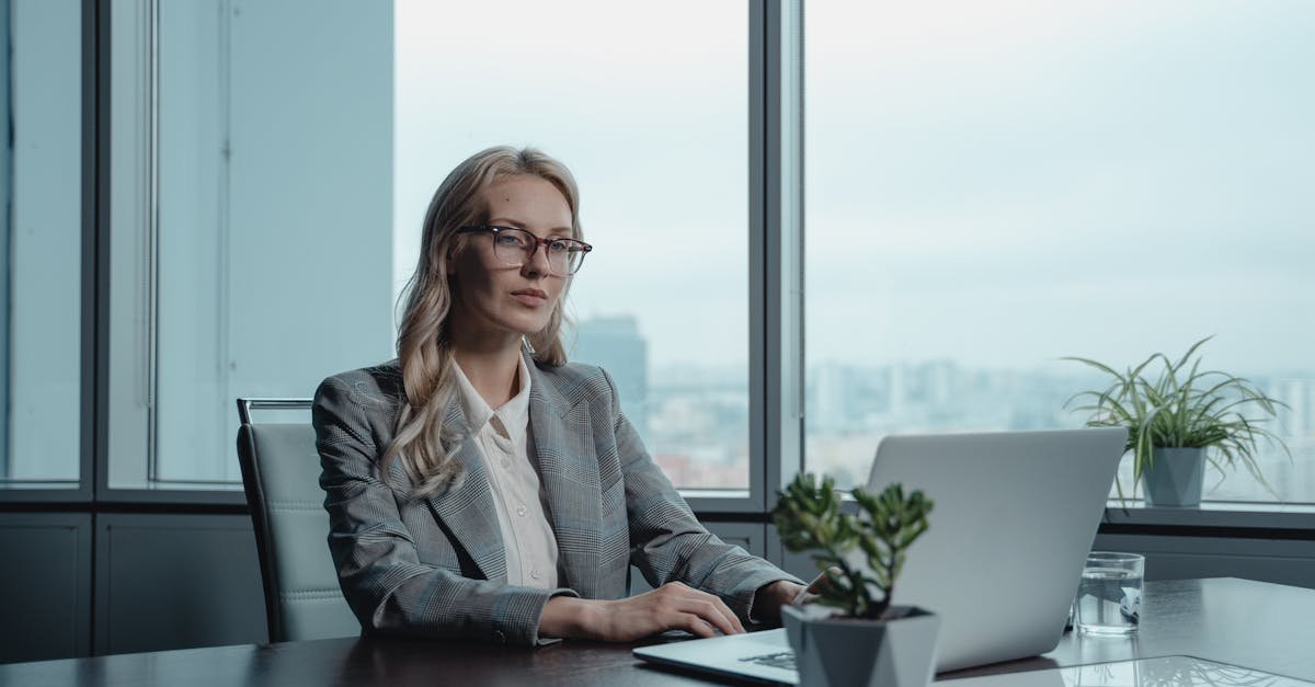 A confident businesswoman in a grey suit working on a laptop in a modern office setting.
