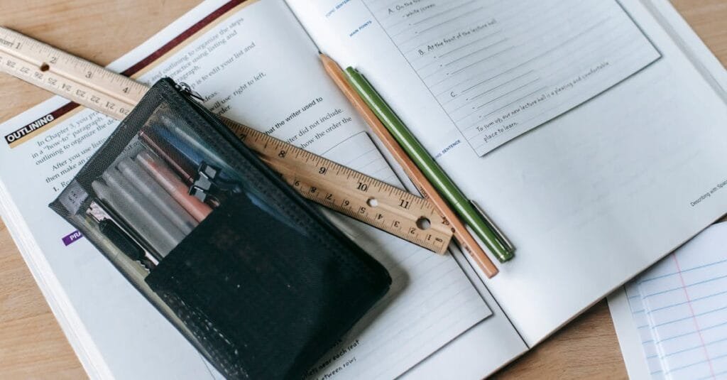 From above of textbook with exercises and ruler with pens near small case on table