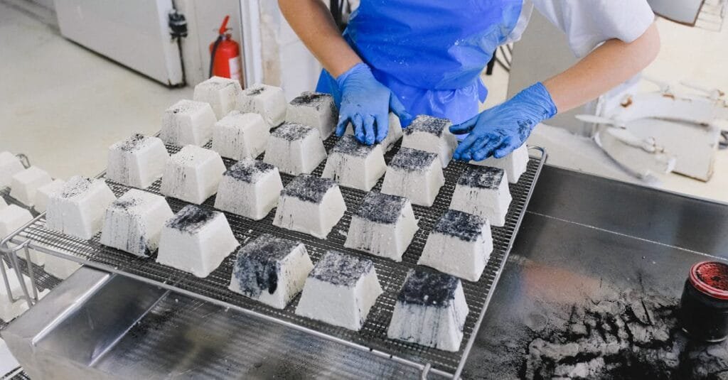 A worker in blue apron and gloves arranges charcoal-coated blocks in a factory setting.
