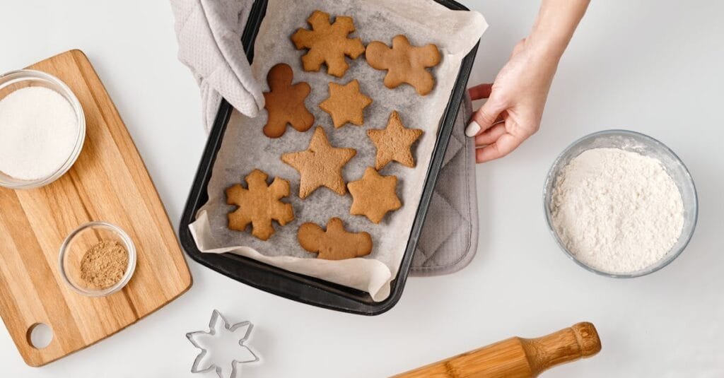 Overhead view of gingerbread cookies on a baking tray with ingredients and cookie cutters.