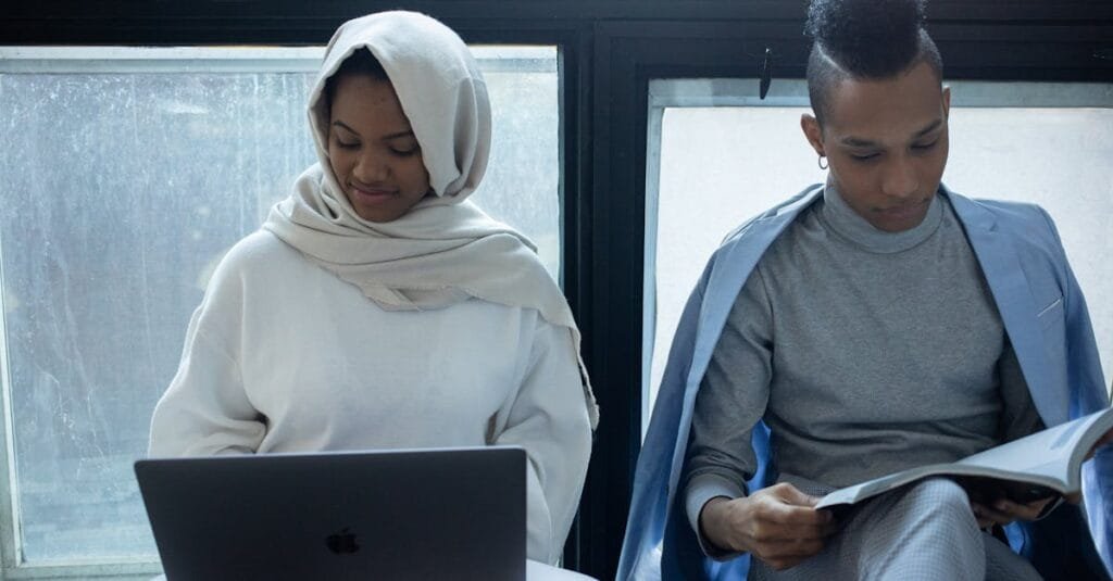 Concentrated African American woman in hijab working on laptop near classmate reading textbook during exam preparation