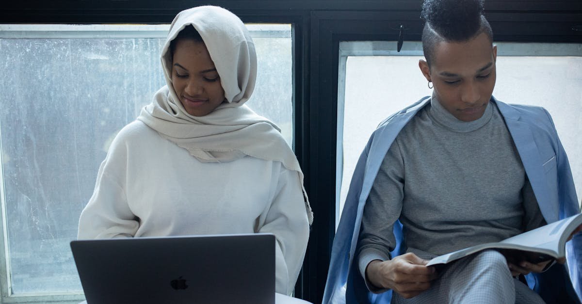 Concentrated African American woman in hijab working on laptop near classmate reading textbook during exam preparation