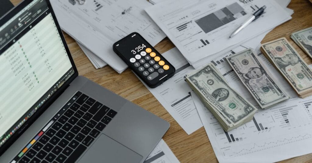 A desk with financial documents, currency, a laptop, and phone calculator.
