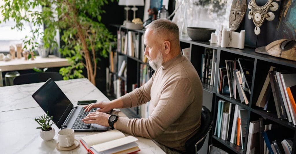 Senior man focused on laptop in a stylish home office, surrounded by books and plants.