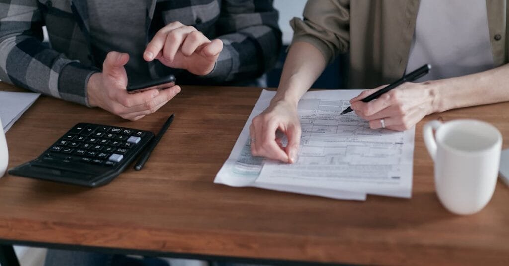 Two people working together on tax forms using a calculator at a wooden desk.