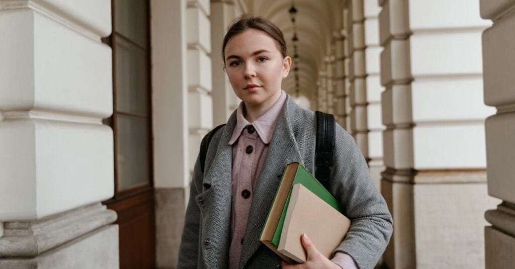 Young woman carrying books in an architectural hallway, dressed for fall weather.