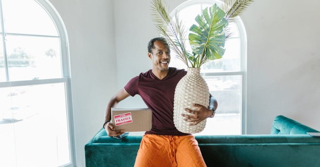 Smiling man holding a fragile box and plant during a home move.
