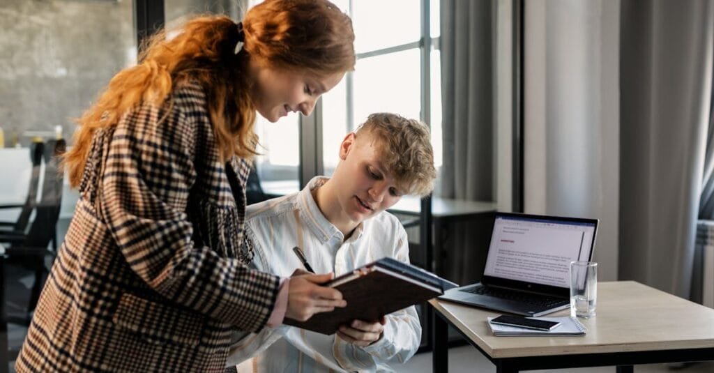 Two colleagues strategizing together in a modern office using a laptop and notepad.