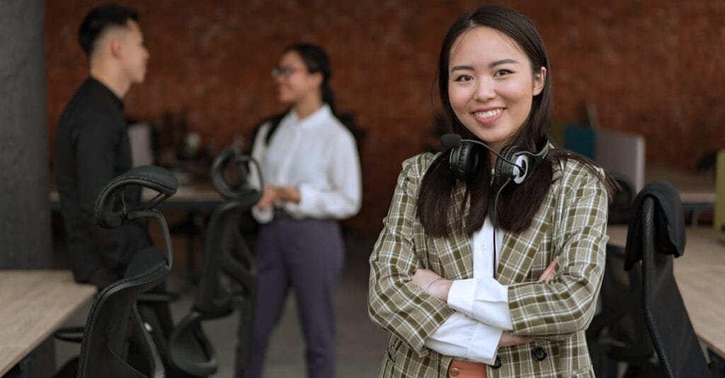 Confident Asian woman with headset and crossed arms in a modern office setting.