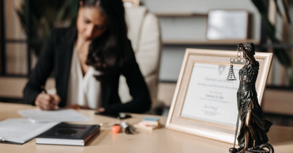 Focused woman writing at desk with Lady Justice figurine and certificate in office.